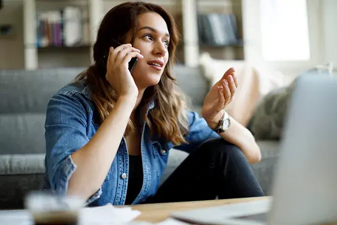 Woman sitting in living room talking on the phone, with a laptop and papers in front of her.