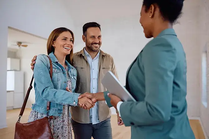 Couple shakes a real estate agents hand while standing inside of an empty living room