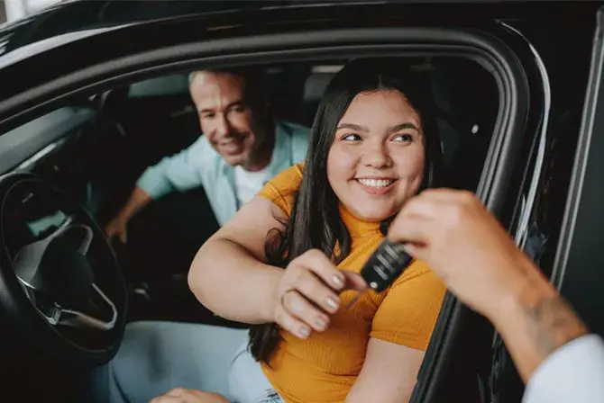 A young woman is seated in the driver’s seat of a car. She smiles as someone hands her the keys to her new car.