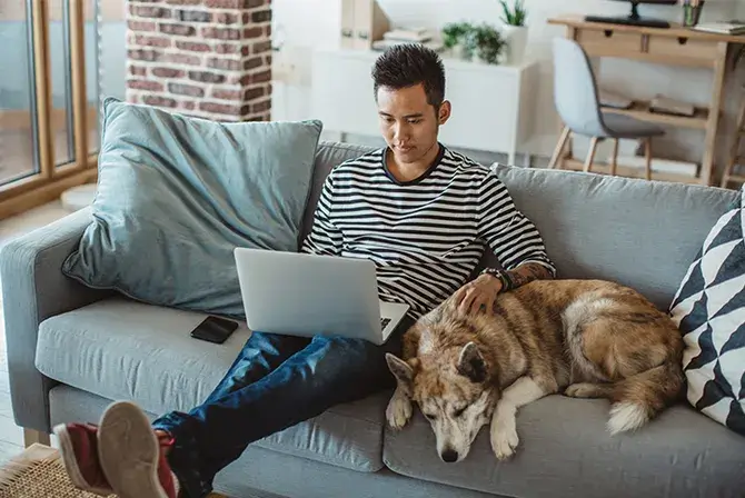A young man sitting on sofa and looking at something on laptop with his pet dog relaxing next to him