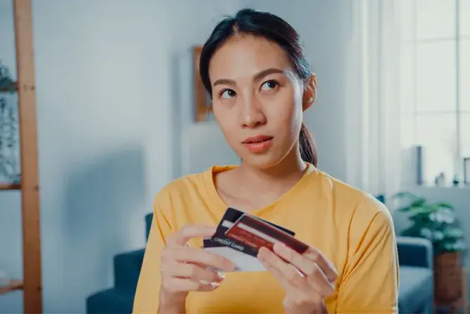 A person in a yellow shirt holding a few credit cards, appearing thoughtful, in a room with soft lighting and modern decor.