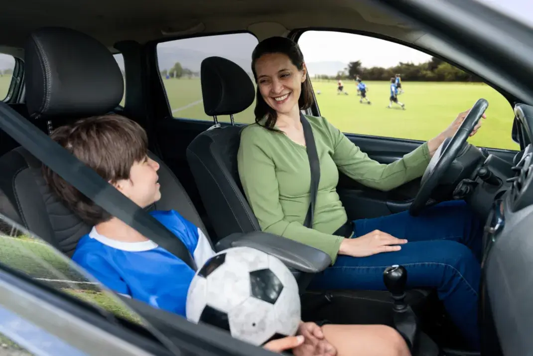 A mom in the driver’s seat of a vehicle faces a child in a sports jersey with a soccer ball in their lap in the passenger seat. A sports field can be seen through the window behind them.