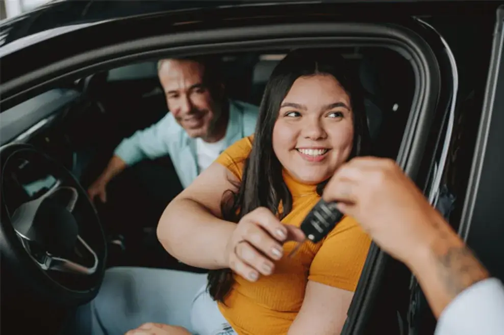 A young woman is seated in the driver’s seat of a car. She smiles as someone hands her the keys to her new car.