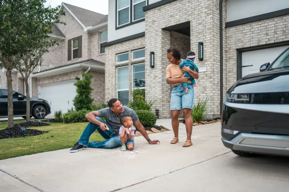A smiling family engages in outdoor fun by their house, with a mother holding her child, as the father draw with chalk with their toddler near their parked car in the driveway.