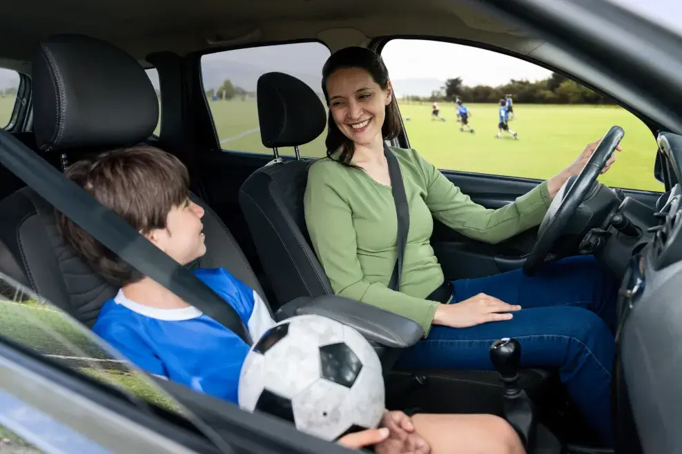A mom in the driver’s seat of a vehicle faces a child in a sports jersey with a soccer ball in their lap in the passenger seat. A sports field can be seen through the window behind them.
