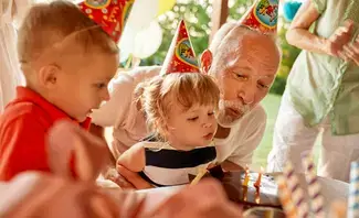 Family celebrating a young child's birthday party outdoors, with an older adult helping the child blow out candles on a cake. Several people, including a child in a party hat, are gathered around the table decorated with party items.