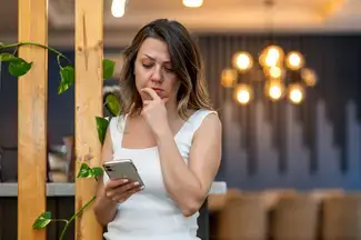 A person looks worried while reading a message on their smartphone in a modern office with hanging lights and plants.