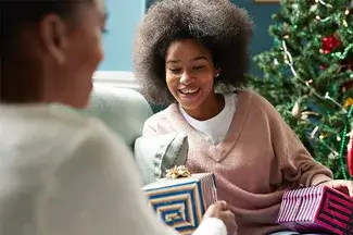 A woman smiles as she receives a wrapped gift from another person who is out of focus in the foreground. A Christmas tree can be seen in the background.