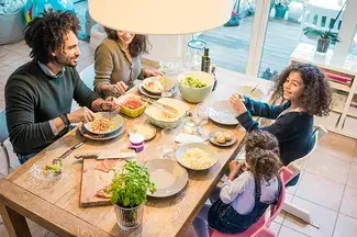 A family is gathered around a dining table enjoying a meal together. The table is filled with dishes, including a salad and a platter of sliced meat. Everyone is engaged in cheerful conversation.