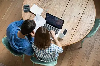 We see two people sitting at a table from an overhead view, using a computer, with paperwork, a phone, and a calculator spread around them.