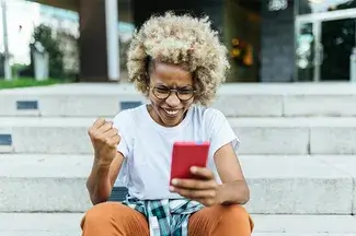 A person sitting on stairs looking at her phone smiles with a celebratory fist pump.