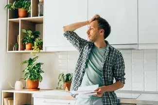 A man in a kitchen surrounded by houseplants holds a document, looking concerned and holding one hand on their forehead.