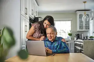 A person points to computer screen while speaking to a person who appears older and could be their parent or loved one.