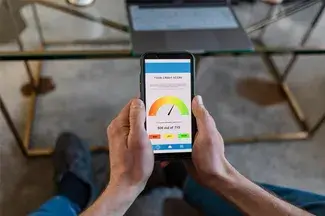 Person using a smartphone displaying a credit score app with a score of 500 out of 775, visible over a glass table with a laptop beside it.