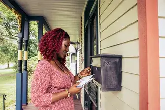 Person checking mail from a mailbox on a house porch, surrounded by greenery.