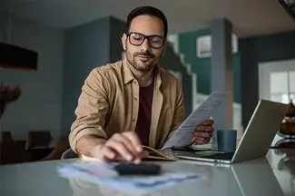 A man sits at a desk with an open lap and smiles confidently as he types into a calculator, holding a piece of paper in his other hand.