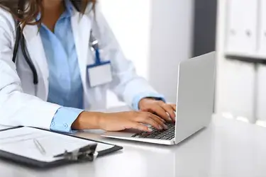 Medical profession sitting at a table with a clipboard off to the side while typing on a computer