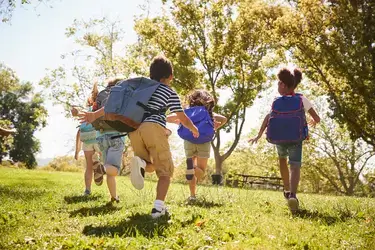 Young children running through a grassy park with backpacks on.