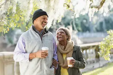 Two people laughing and enjoying coffee together outdoors in a park with tree foliage in the background.