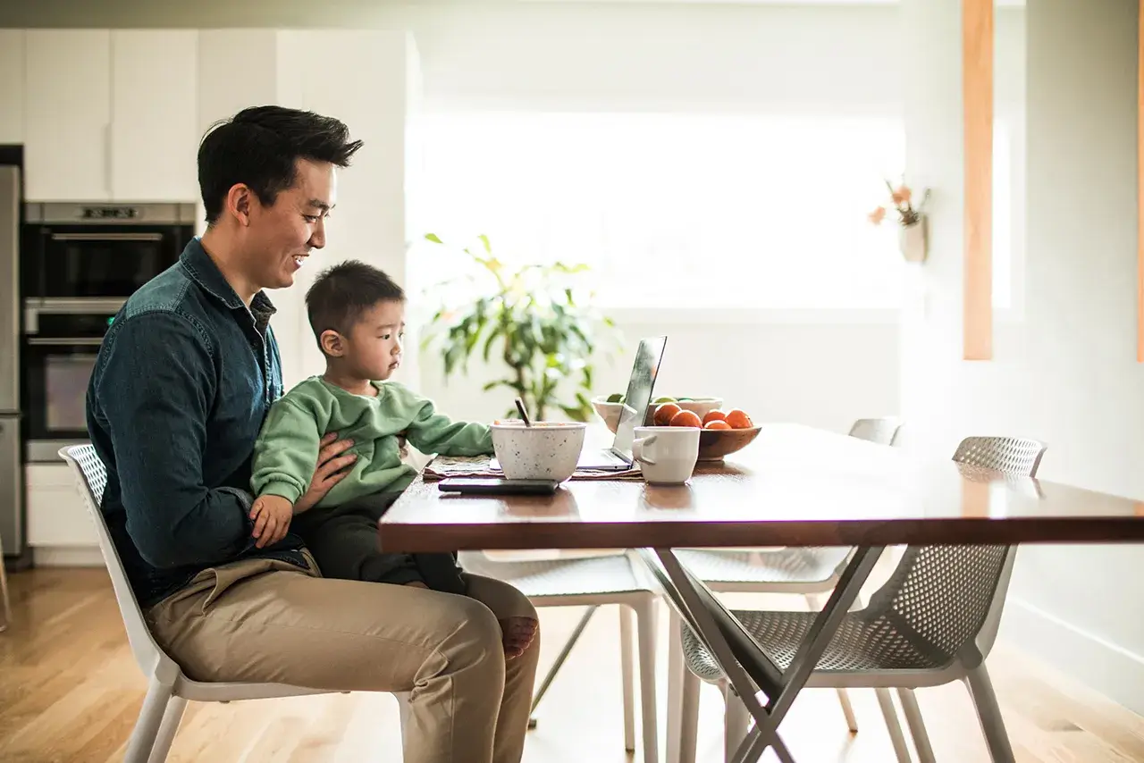 Man sits with young child at a kitchen table, smiling and looking at laptop 